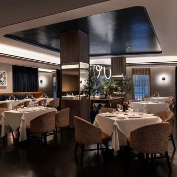 A dimly lit restaurant dining area with several tables set with white tablecloths and brown chairs.