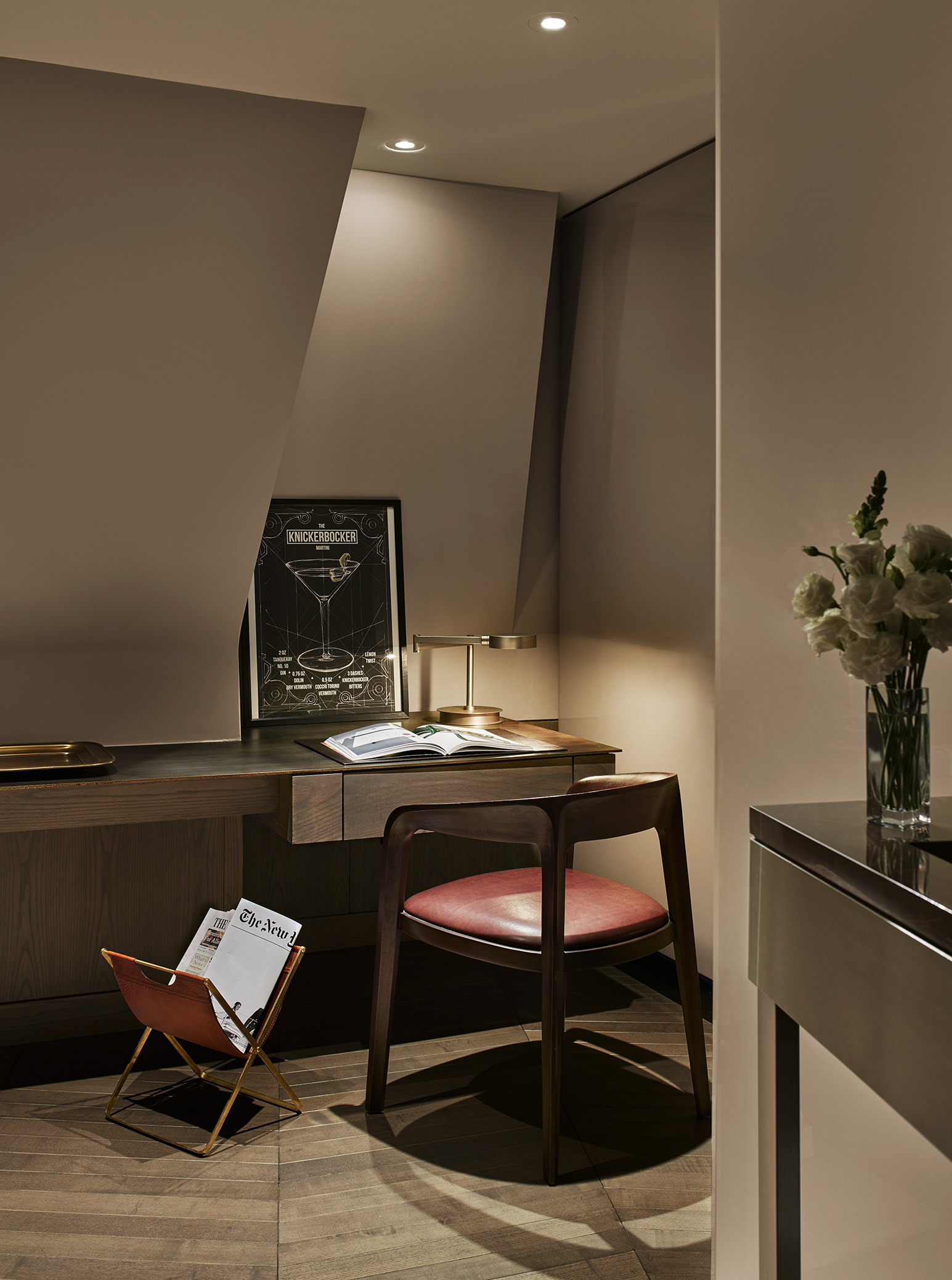 A hotel room desk with a chair, lamp, flowers, and a poster.