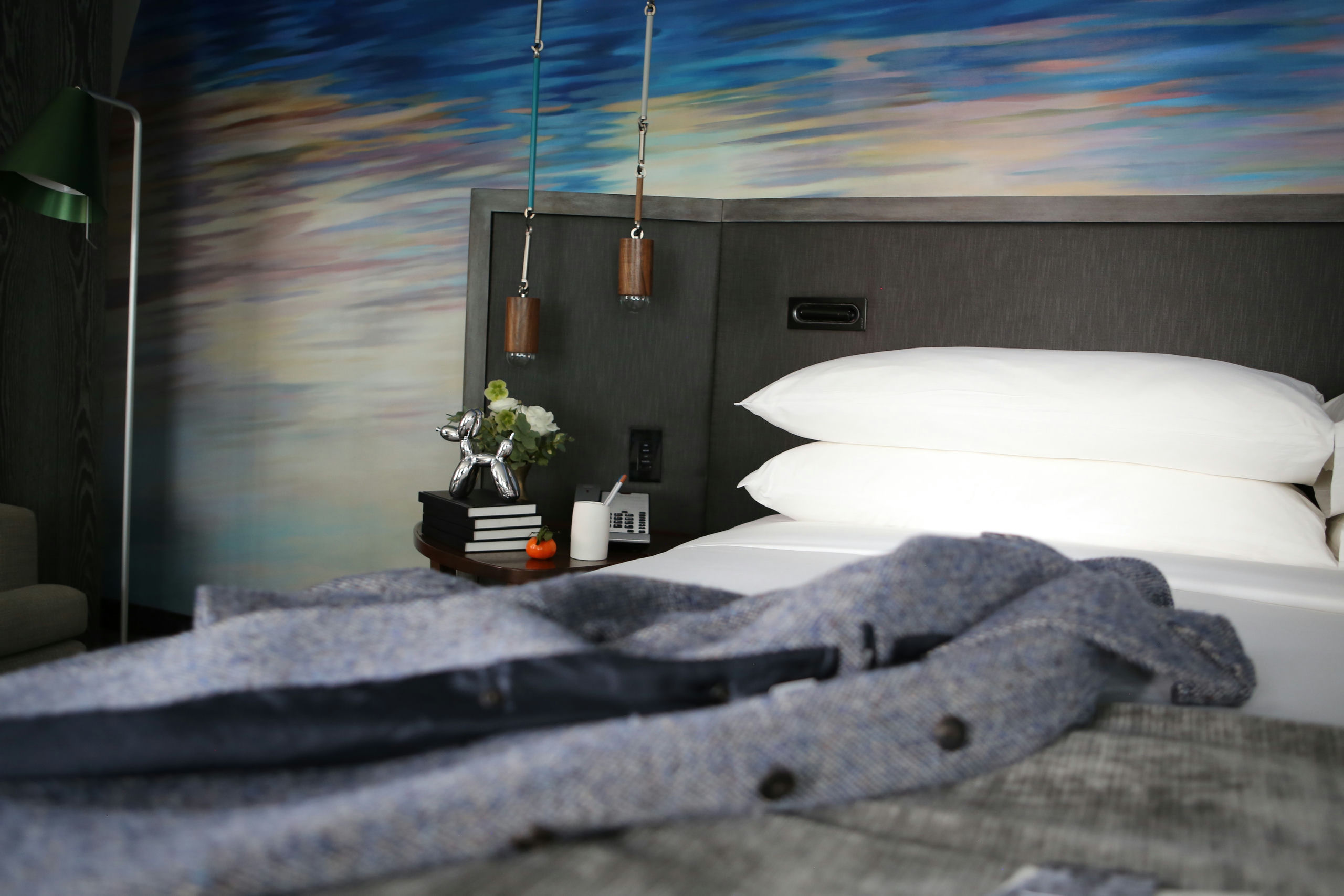 A close-up view of a bed with white bedding and pillows, a nightstand with a lamp, books, and flowers.