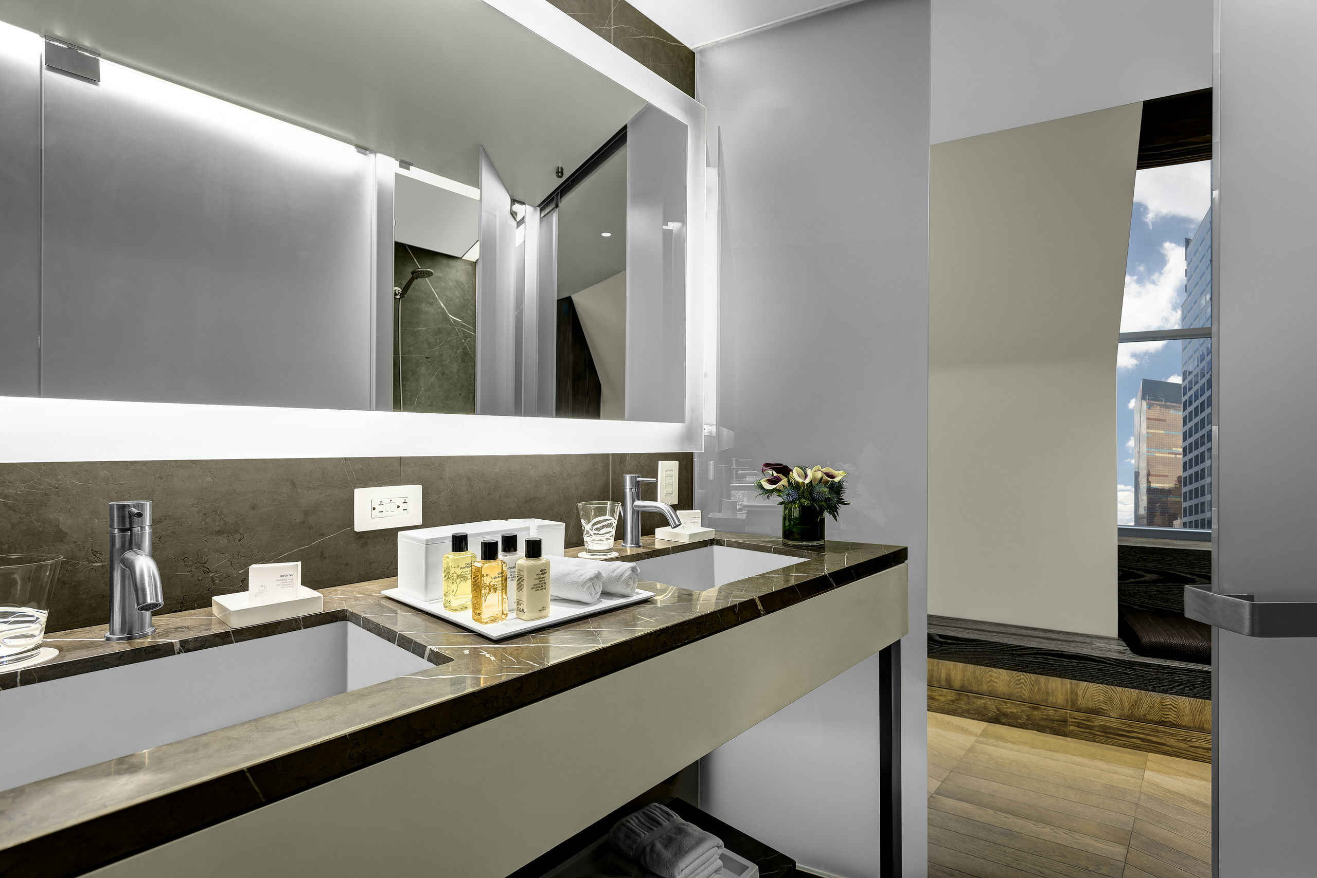 A modern hotel bathroom vanity with a large lit mirror, marble countertop, and amenities.
