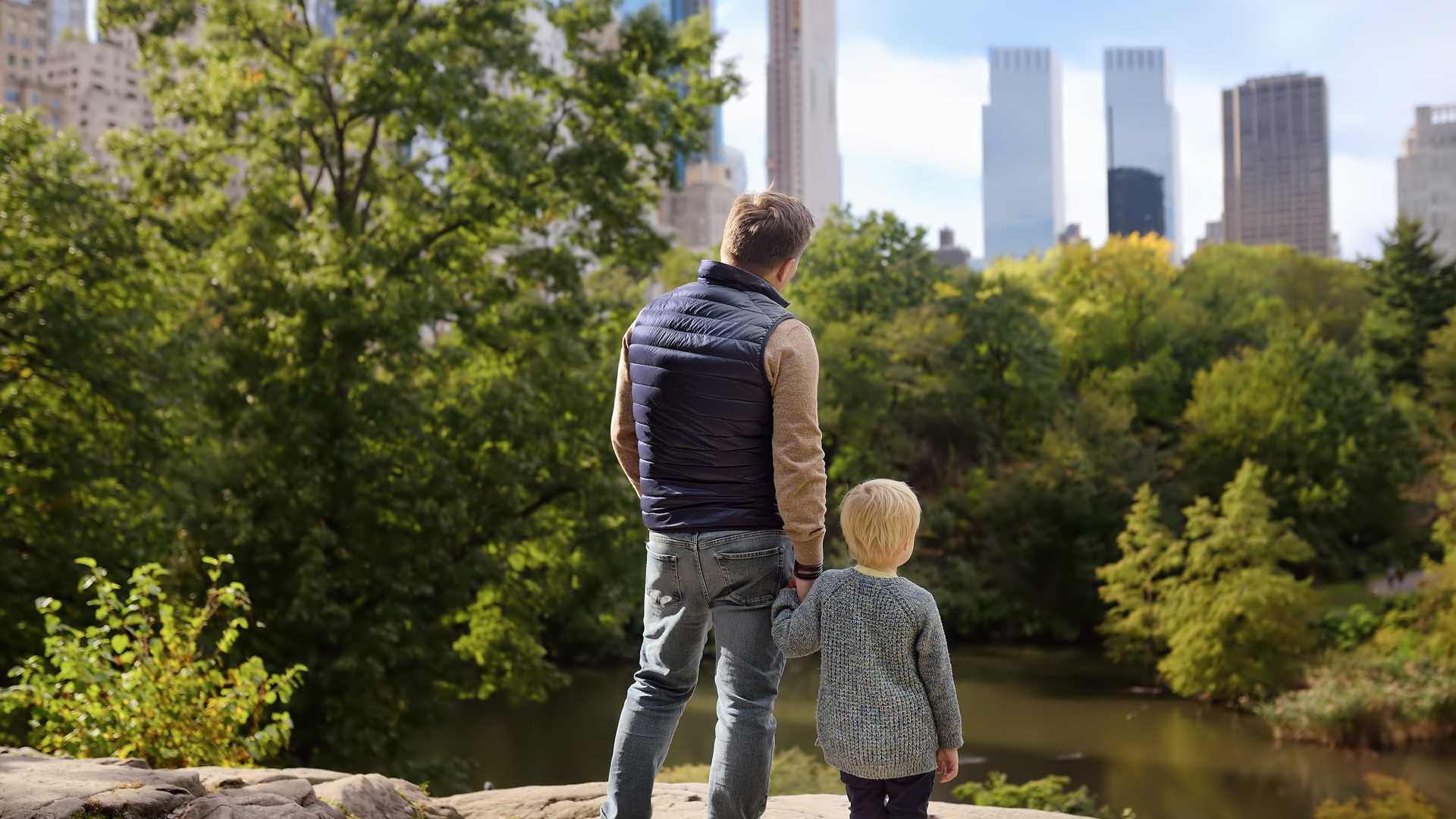 man and child looking at a pond and the city buildings of NYC
