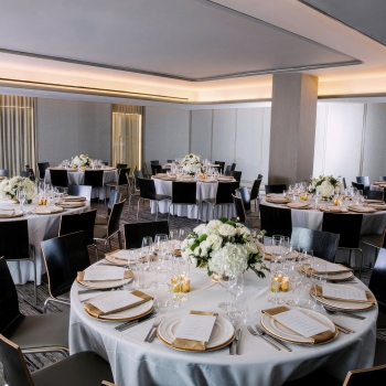 A large, elegantly set dining room with round tables, white tablecloths, black chairs, and floral centerpieces.