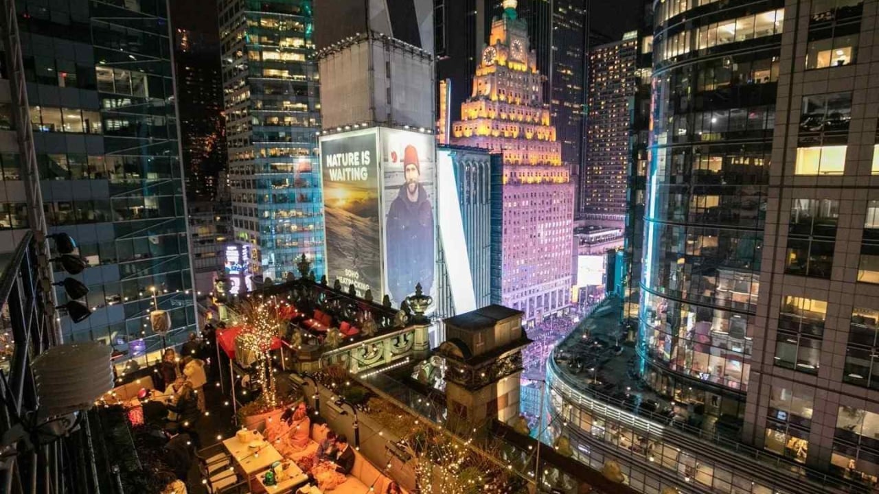 A nighttime view of a city street with tall buildings and a rooftop restaurant or bar