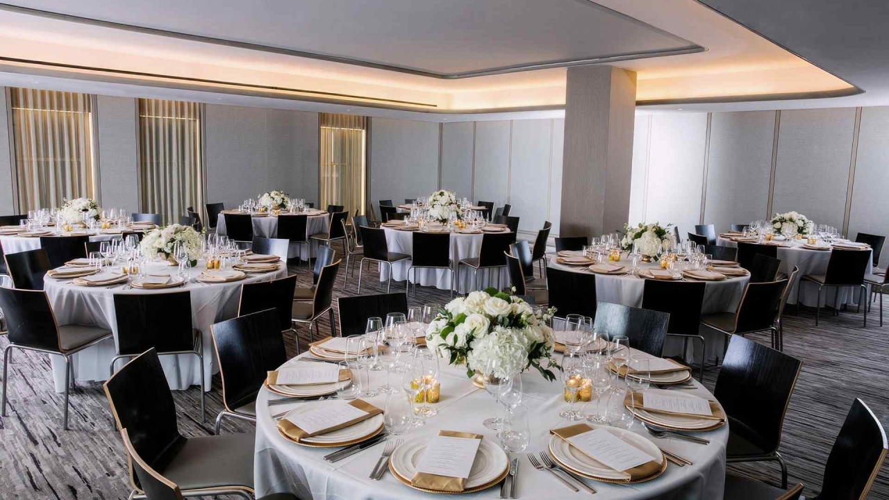 A large, elegantly set dining room with round tables, white tablecloths, and black chairs, adorned with white flowers and candlelight.