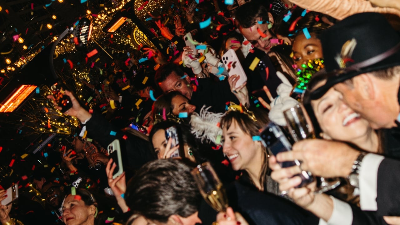 A crowded bar scene with people celebrating, surrounded by confetti, and some holding champagne and phones.